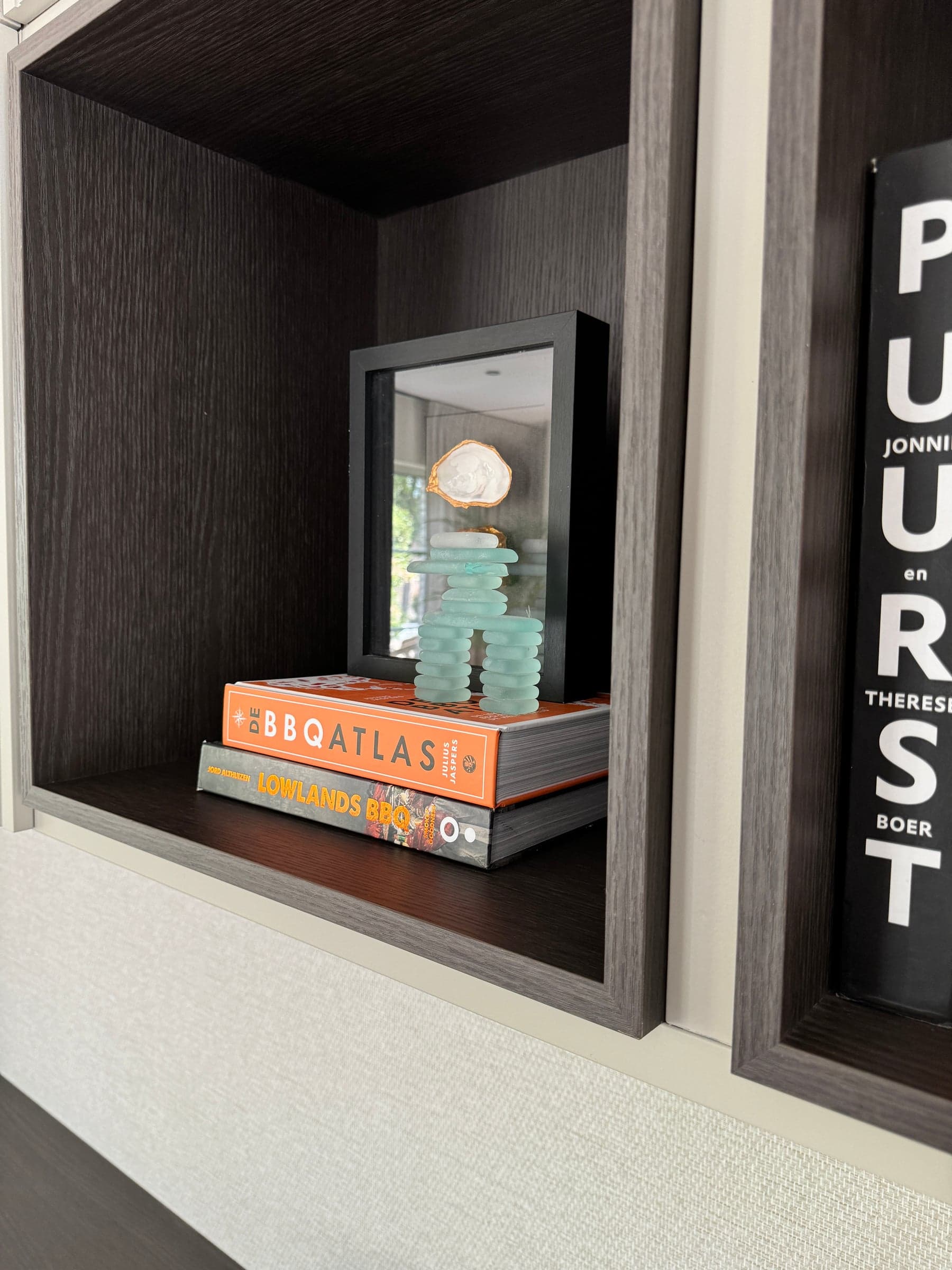 Stylingdetail van een donkere eiken wandkast met designboeken en een schelp in een lijstje in een woonhuis in Best
