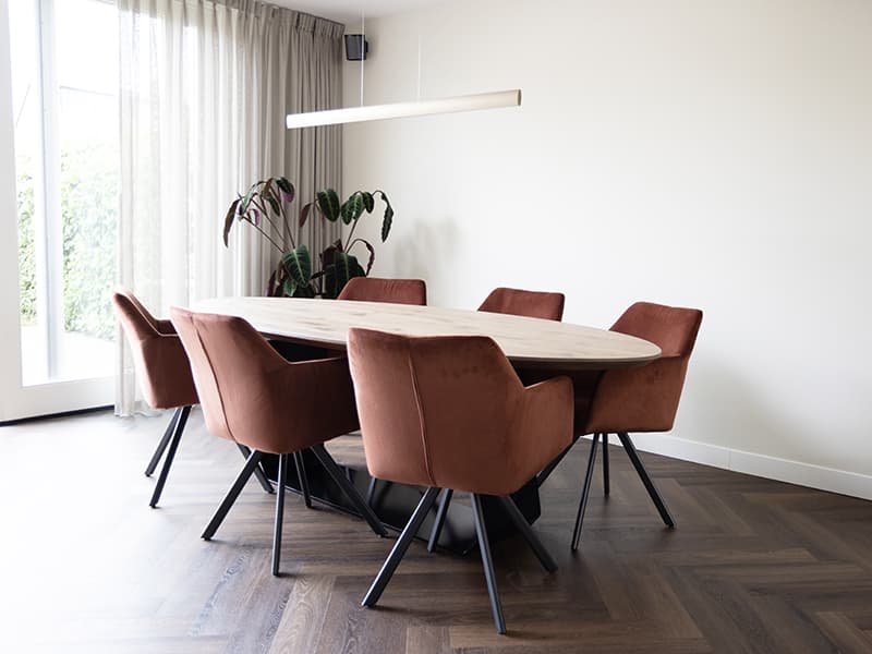 Ovale eikenhouten eettafel met terracotta velours stoelen op een visgraatvloer in een woonhuis in Oisterwijk