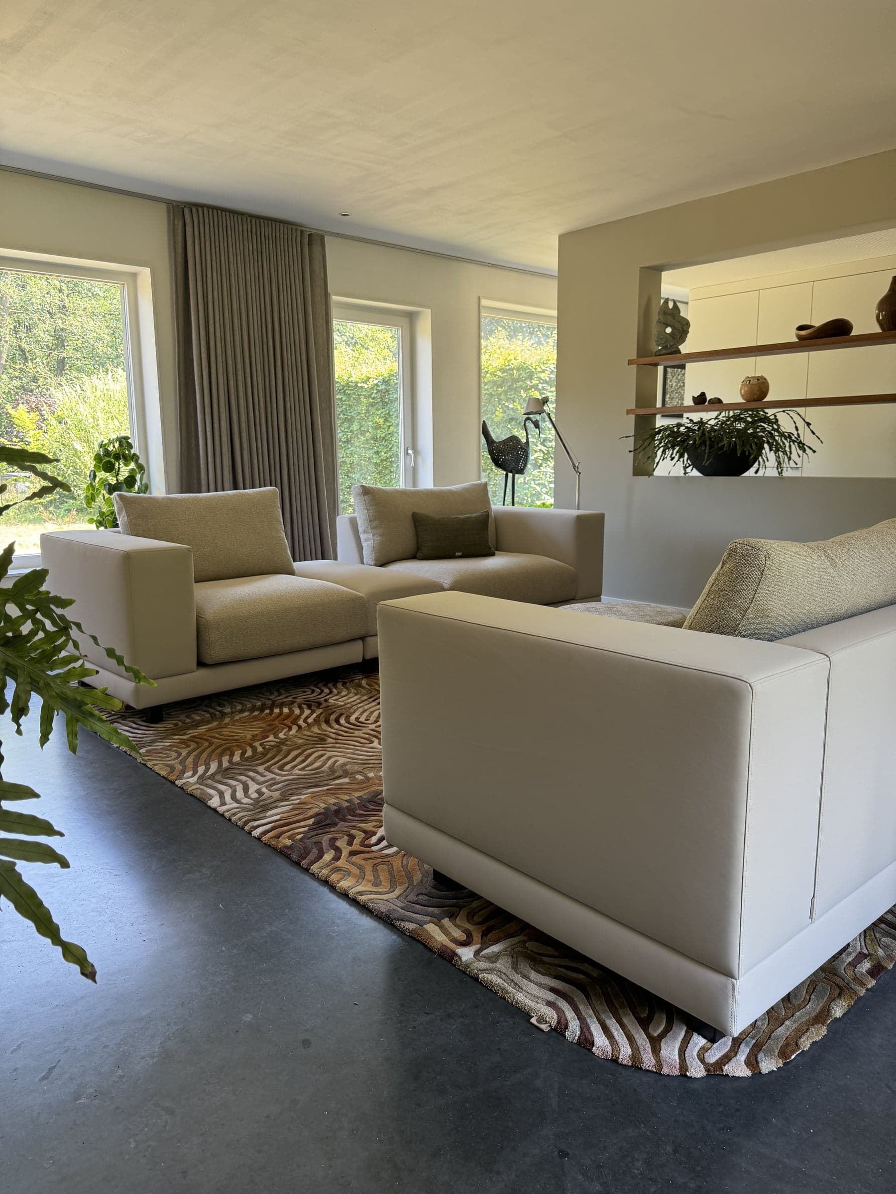 Woonkamer met zandkleurige bank en fauteuils, een gedessineerd vloerkleed en grote raampartij in een woonhuis in Oisterwijk