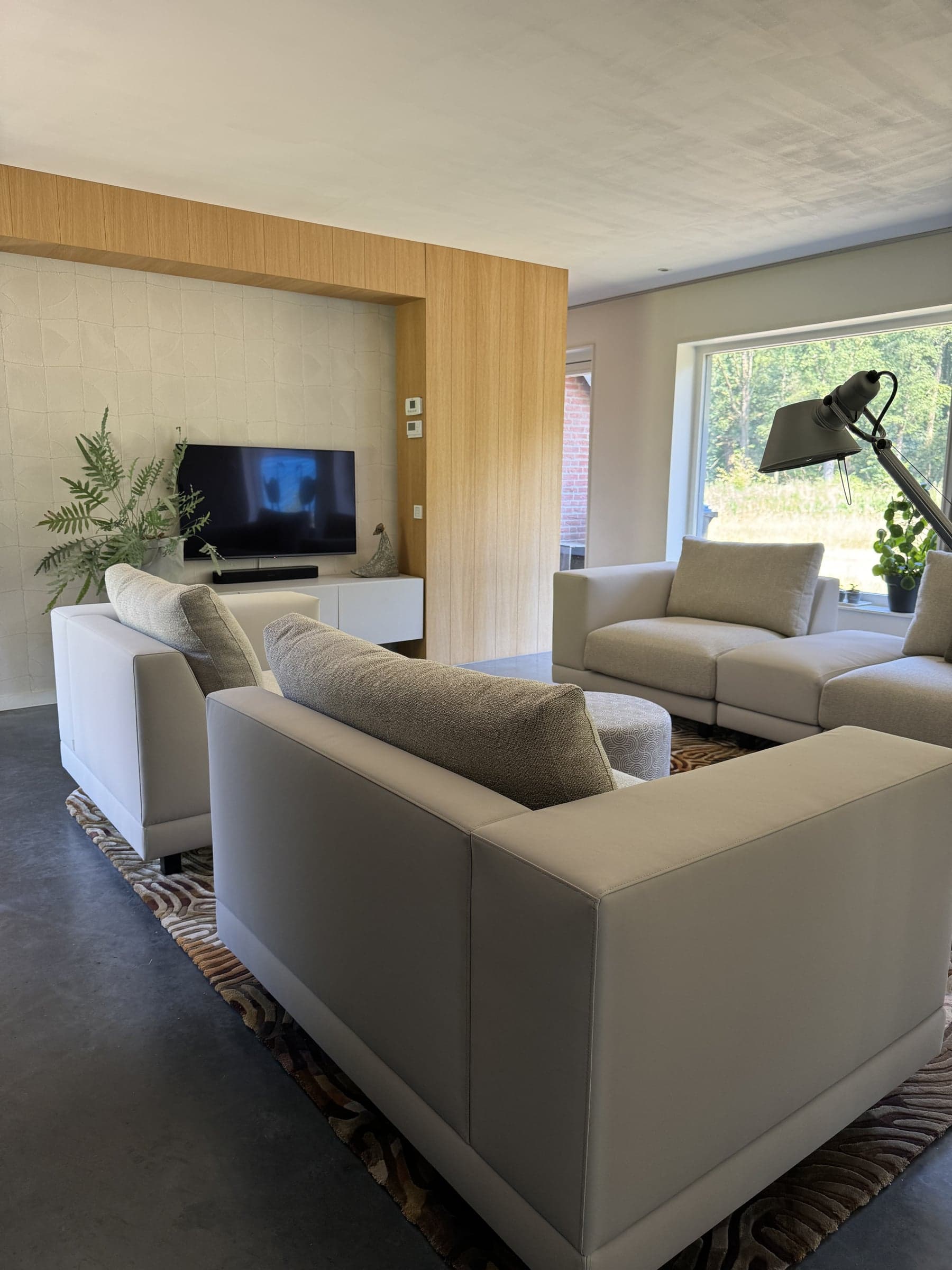 Woonkamer met zandkleurige banken, houten wandpaneel en tv op een betonvloer in een woonhuis in Oisterwijk